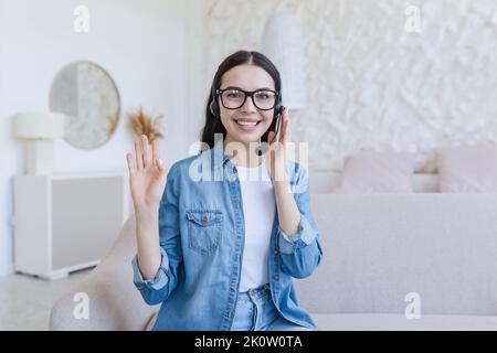 Webinar. Junge, schöne Geschäftsfrau mit Brille und Kopfhörer sitzt zu Hause auf dem Sofa und führt ein Webinar, Online-Schulungen vor der Kamera, berät, erzählt, Gesten mit den Händen, lächelt. Stockfoto
