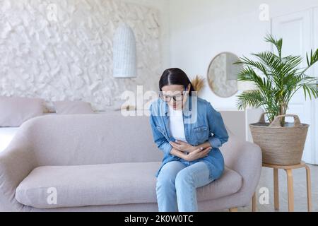 Bauchschmerzen. Junge schöne Frau in Brille und Jeans sitzt auf dem Sofa zu Hause und hält ihren Bauch. Fühlt sich scharfe Schmerzen, Vergiftung, fühlt sich schlecht Stockfoto