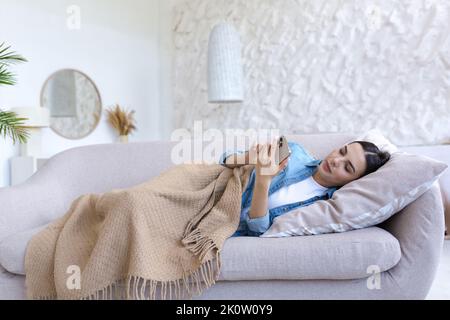 Eine junge, schöne Frau liegt zu Hause auf dem Sofa, hält das Telefon in den Händen, schreibt eine Nachricht, wählt. Bedeckt mit einer beigen Decke, in Denim-Kleidung. Er ist krank, macht ein Nickerchen, arbeitet online von zu Hause aus Stockfoto