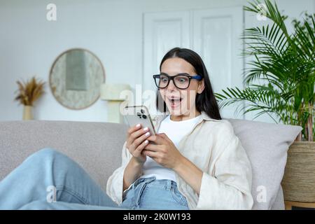 Glückliche junge Frau in Brille mit Mobiltelefon, feiern unerwarteten Sieg im Spiel, gewonnen. Zu Hause auf dem Sofa sitzen, aufgeregt, zufrieden. Stockfoto