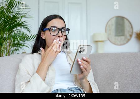 Schockierte junge, schöne Frau mit Brille schaut auf das Telefon, bedeckt ihren Mund mit ihren Händen. Hat eine Nachricht erhalten, liest die Nachrichten, schaut sich ein Video an. Zu Hause auf der Couch sitzen. Stockfoto