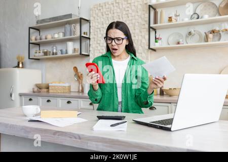 Verwirrt und nervös junge schöne Frau in Brille sitzt zu Hause in der Küche mit einem Laptop und berechnet ihr Familienbudget. Er hält Rechnungen, Dokumente und ein Telefon in den Händen. Stockfoto