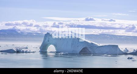 Massiver Eisberg mit großen Bögen vor dem Hintergrund schneebedeckter Berge in Qaqortoq, Grönland Stockfoto