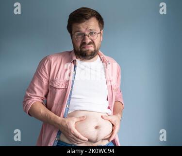 Unglücklich, unzufrieden übergewichtigen Mann, hob T-Shirt und hält überschüssiges Bauchfett mit den Händen. Gesundes Lifestyle-Konzept. Stockfoto