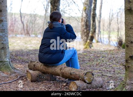 Die junge Frau sitzt auf einer Holzbank im Wald und trinkt Kaffee. Alter Holzsitz im Park. Stockfoto