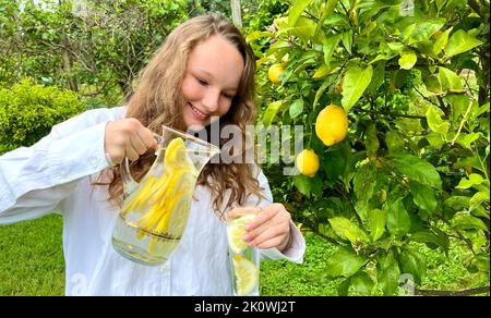 Es ist nur ein Zitronenparadies, ein Teenager-Mädchen steht vor dem Hintergrund eines Zitronenbaums mit Zitronen und trinkt Limonade aus einem Glas in ihrem anderen h Stockfoto