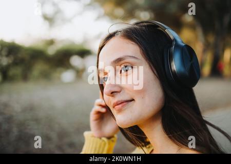 Nahaufnahme eines Teenagers in gelbem Pullover, das im Park mit einem Kopfhörer Musik auf dem Smartphone spielt Stockfoto