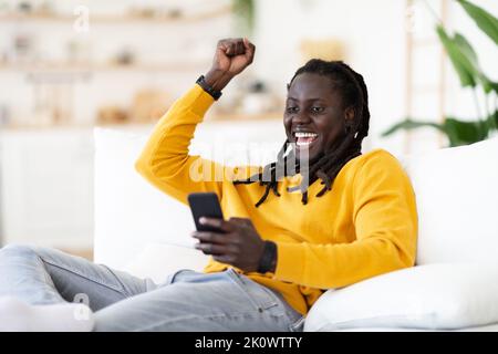 Porträt Des Fröhlichen Afroamerikanischen Guy Mit Smartphone Feiert Erfolg Zu Hause Stockfoto