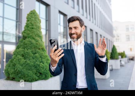Fröhlicher, selbstbewusster, hübscher, junger kaukasischer Manager mit Bart im Anzug winkt der Hand, führt einen Videoanruf Stockfoto