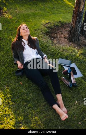 Ruhige, schöne kaukasische Geschäftsfrau, die mit geschlossenen Augen auf Gras liegt und die frische Morgenluft genießt. Draufsicht. Eine fokussierte freiberufliche Mitarbeiterin arbeitet Stockfoto