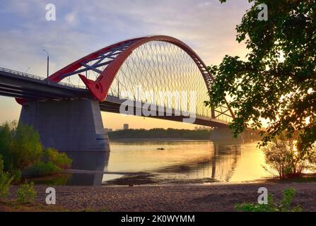 Eine große Bogenbrücke im Morgengrauen. Bugrinsky Brücke über den ob Fluss zwischen den Ästen von Bäumen. Nowosibirsk, Sibirien, Russland, 2022 Stockfoto