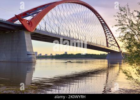 Eine große Bogenbrücke im Morgengrauen. Bugrinsky Brücke über den ob Fluss zwischen den Ästen von Bäumen. Nowosibirsk, Sibirien, Russland, 2022 Stockfoto