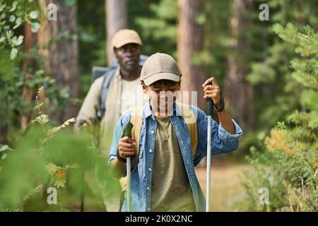 Lächelnder jugendlicher Junge mit Trekkingstöcken, der vor seinem Großvater auf einem Wanderpfad im Pinienwald unterwegs ist Stockfoto