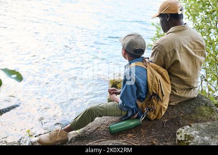 Rückansicht des afroamerikanischen Jungen mit Rucksack und seines Großvaters, der am Wasser sitzt und sich während der Wochenendwanderung ausruhen kann Stockfoto