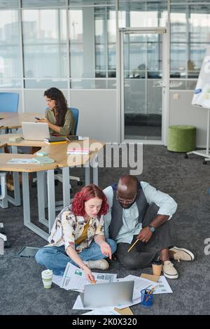 Junge Geschäftsfrau zeigt während der Präsentation neuer Projektpunkte auf den Laptop-Bildschirm und diskutiert diese mit einem afroamerikanischen Mann Stockfoto