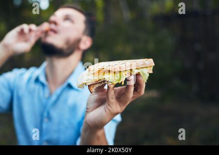 Ein junger Mann isst ein Sandwich in der Natur. Vorderansicht. Stockfoto