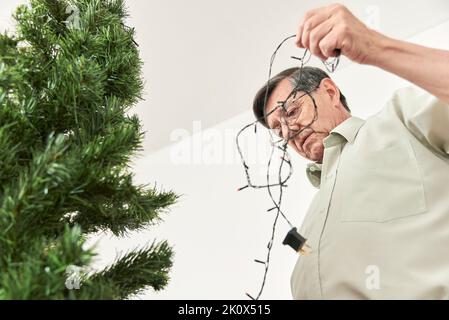 Bedenken bezüglich der Nutzung elektrischer Energie während der Feiertage. Hispanischer älterer Mann, der eine Lichterkette ausstreckt, um in seinem Haus einen Weihnachtsbaum anzuziehen Stockfoto