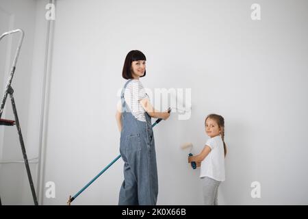 Reparatur in der Wohnung. Rückansicht der glücklichen Familie von Mutter und kleinen niedlichen Kind Tochter malt die Wand mit weißer Farbe. Stockfoto