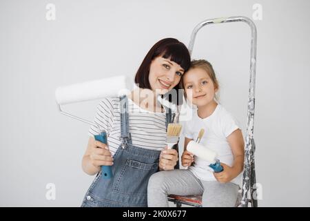 Eine fröhliche glückliche Familie renoviert eine erworbene Wohnung. Die Mutter umarmt ihre Tochter, sitzt auf einer Leiter, mit Freude. Jeder hält eine Farbrolle und Pinsel, um die Wände zu malen. Stockfoto