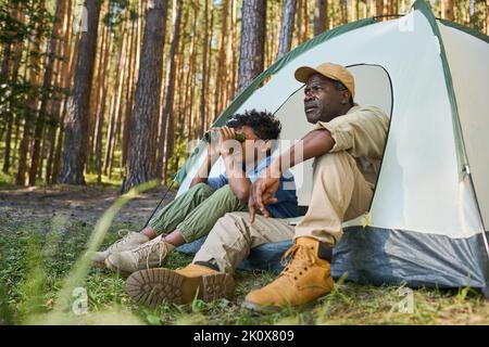 Süßer afroamerikanischer Junge, der durch ein Fernglas schaut, während er im Zelt neben seinem Großvater in Casualwear während der Wanderung im Wald sitzt Stockfoto