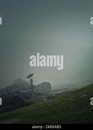 Mann mit Regenschirm, der in einem düsteren Wetter mit dichtem Nebel einen Berg besteigen kann. Moody Wanderszene mit einem Wanderer, der auf den Klippen des nebligen Stockfoto