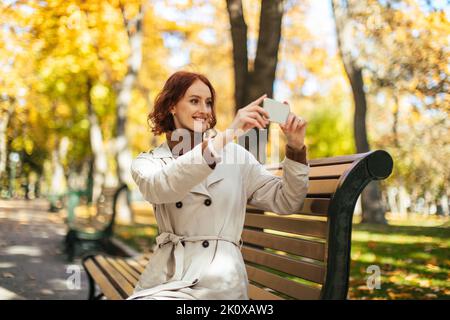 Lächelnd sitzt das hübsche kaukasische, tausendjährige rothaarige Weibchen im Regenmantel auf der Bank und fotografiert, genießt den Herbst Stockfoto
