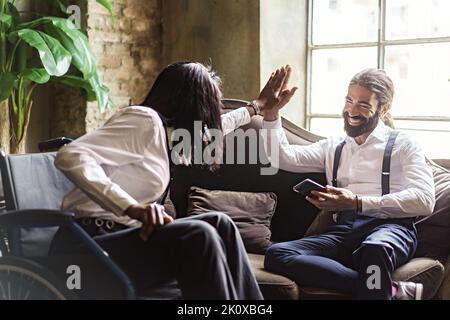 Eine afroamerikanische Geschäftsfrau mit Behinderungen sitzt auf einem Rollstuhl und nimmt mit ihren Kollegen im Büro die fünf höchsten Punkte ein - der Lebensstil der Geschäftsleute Stockfoto