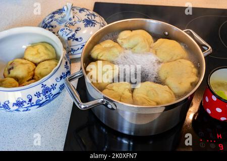 Süße Fruchtknödel, die in kochendem Wasser in einem Topf auf einem heißen Keramikkochfeld schwimmen, gekochter Knödel in einer dekorativen Schüssel, Deckel, geschmolzene Butter in kleinen roten Punkten Stockfoto