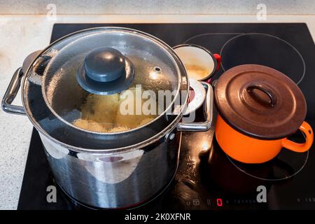 Gekochte süße Pflaumenknödel im Topf mit Deckel auf heißem Keramikkochfeld, geschmolzene Butter im rot gepunkteten Topf, orangefarbener Topf, Nahaufnahme. Stockfoto