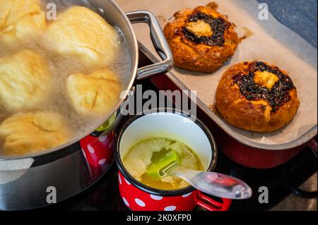 Süße Fruchtknödel, die in kochendem Wasser im Topf schwimmen, geschmolzene Butter im Topf auf einem Cerankochfeld, zwei Obstkuchen auf dem Küchentisch. Stockfoto