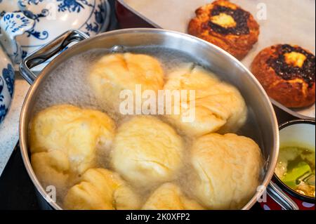 Süße Fruchtknödel, die in kochendem Wasser im Topf auf einem Keramikkochfeld schwimmen, geschmolzene Butter in einem kleinen Topf, 2 Futi-Kuchen auf dem Tisch, Nahaufnahme. Vorbereitungsknödel Stockfoto