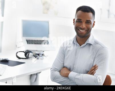 Die Arbeit hält mich in Schuss. Ein hübscher junger afroamerikanischer Geschäftsmann sitzt mit gefalteten Armen an seinem Schreibtisch. Stockfoto