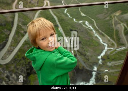 Niedliches Kind, das auf die Kamera schaut, während es auf dem Aussichtspunkt der Trollstigen Straße steht, der berühmten Straße im Sommer in Norwegen Stockfoto