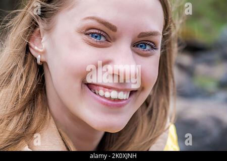 Porträt einer jungen attraktiven blauäugigen Frau, die die Kamera anschaut und in einer gelben Jacke lächelt. Selektiver Fokus Stockfoto