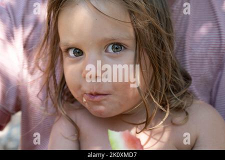 Porträt eines niedlichen Babys. Das Kind isst eine Wassermelone und schaut auf die Kamera. Lifestyle. Offen. Das Konzept eines aktiven Familienurlaubs. Vorderansicht. Nahaufnahme. Stockfoto