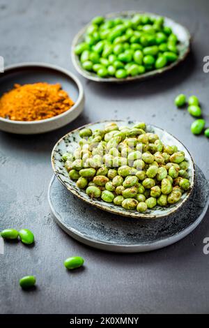 Geröstete Edamame-Bohnen als Snack mit Gewürzen und rohen Bohnen im Hintergrund. Gesunde Alternative zu Kartoffelchips Stockfoto