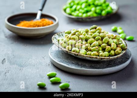 Geröstete Edamame-Bohnen als Snack mit Gewürzen und rohen Bohnen im Hintergrund. Gesunde Alternative zu Kartoffelchips Stockfoto