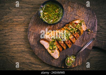 Hausgemachtes gegrilltes Schweinesteak mit Chimichurri-verde-Sauce auf Schneidebrett auf dunklem Hintergrund Stockfoto