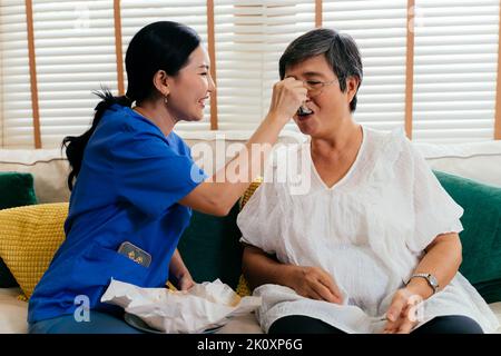 Asiatische Frau in blauen Peelings füttern ältere weibliche Patientin, während sie auf dem Sofa im hellen Raum zu Hause sitzt Stockfoto
