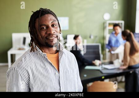 Porträt eines afrikanischen Mannes, der die Kamera anlächelt, während er mit seinen Kollegen im Büro arbeitet Stockfoto