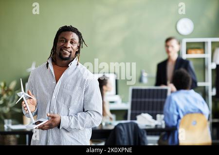 Porträt eines afrikanischen Ingenieurs mit Windmühlenmodell, das die Kamera anlächelt, während er im Büro steht Stockfoto