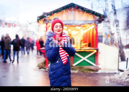 Junge Frau trinkt Punsch auf dem weihnachtsmarkt. Nettes schönes Mädchen in rotem Hut steht auf der Straße und trinkt heißen Wein oder Tee oder Kakao, heißen Kaffee im Freien im Winter Schneetag. Weihnachtsbeleuchtung Stockfoto