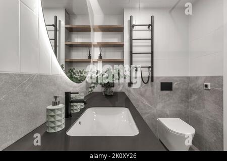 Moderne, minimalistische Einrichtung im Badezimmer mit grauen Steinfliesen, schwarzen Möbeln, Eukalyptus in Glasvase, rundem Spiegel. Ästhetisch einfaches Interio Stockfoto