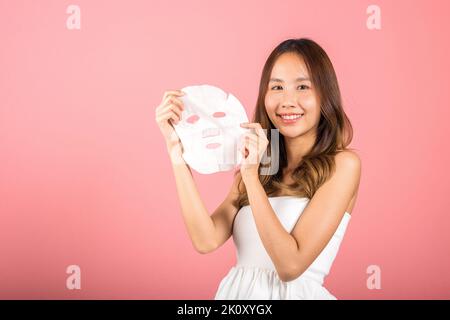 Gesundheitszentrum. Portrait schöne Frau lächelt hält Gesichtsmaske, Studio Shot isoliert auf blauem Hintergrund, asiatische Frau hält reinigende Maske Stockfoto