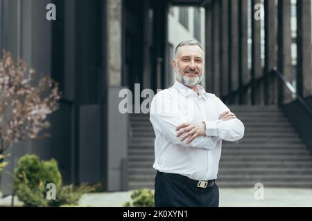 Porträt eines älteren Mannes, eines Lehrers, eines Lehrers, der in einem weißen Hemd vor dem Hintergrund eines modernen Gebäudes einer Bildungseinrichtung steht. Grillen, schauen Sie sich die Kamera an Stockfoto