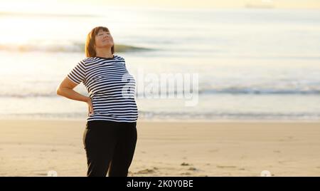 Glückliche Frau im mittleren Alter arbeitet an Übungen an einem Sandstrand am Meer bei Sonnenaufgang oder Sonnenuntergang. Weiblich 50 s kümmert sich um Körper und entspannend. Sport, Stockfoto