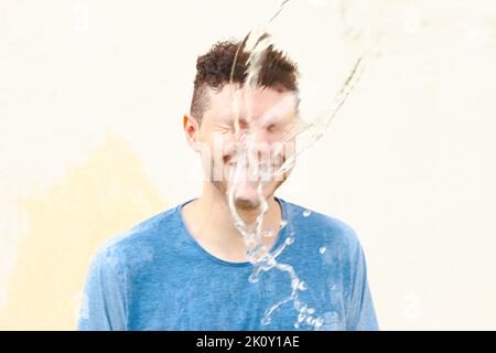 Nass lächelnder junger kaukasischer Mann in blauem T-Shirt mit geschlossenen Augen im Wasserspritzer, Wasserstrahl auf gelbem Hintergrund. Stress, Schock. Vorderansicht, Port Stockfoto