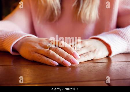 Weibliche Hände mit langen Nägeln, rosa Maniküre und Ringe Schmuck auf Holztisch. Nahaufnahme, Kopierraum, Wartekonzept. Stockfoto