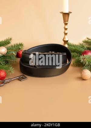 Aufgerollter schwarzer modischer Herren Ledergürtel mit automatischer Schnalle, Weihnachtstanne, glänzenden Kugeln, goldenem Kerzenständer und Schlüssel auf goldenem Hintergrund. Stockfoto