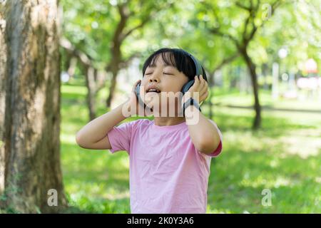 Portrait von Kind Mädchen hört Musik mit modernen Kopfhörern im Park im Freien. Fröhliches Kind, das mit Kopfhörern über den wi-Speed-Kopfhörer Rhythmen hört Stockfoto